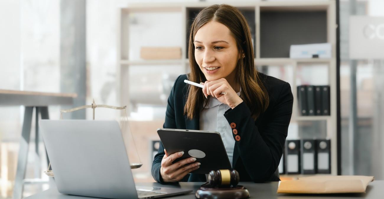 Ein kleines, helles Büro. Im Hintergrund ein Regal mit Akten. Davor am Schreibtisch sitzt eine junge Frau. Vor ihr liegt ein Richterhammer. Sie studiert gleichzeitig am Notebook und am Tablet Infos zu Verwaltungssoftware für regulierte Branchen.