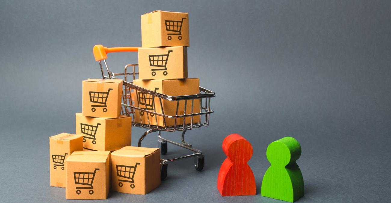 Ein stilisiertes Bild mit grauem Hintergund: Im Vordergrund stehen eine rote und eine grüne hölzerne Spielfigur neben einem kleinen Einkaufswagen, der mit Paketen überhäuft ist. Das Symbolisiert eine Warenwirtschaftssystem für den Einzelhandel