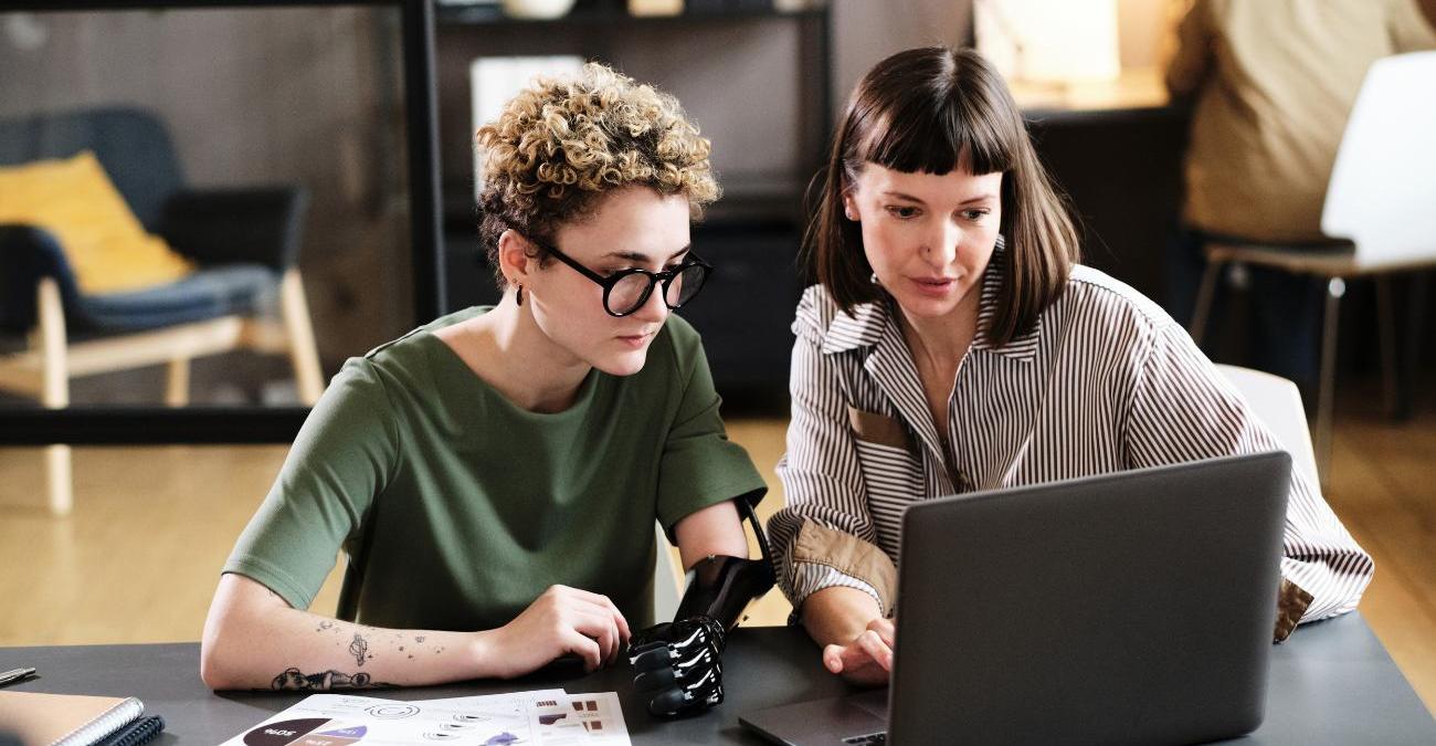 Ein modernes Office. Zwei Mitarbeiterinnen sitzen zusammen an einem Notebook. Sie arbeiten harmonisch. Eine von ihnen trägt eine Armprothese. Gemeinsam arbeiten sie daran, WCAG 2.2 im Büro umzusetzen.