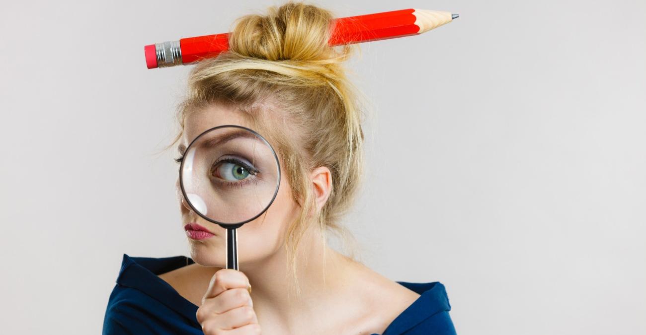 Eine junge Frau trägt die Haare in einem Dutt um einen überdimensionalen roten Bleistifft gewunden. Sie blickt durch eine große Lupe dem Leser entgegen. Ihr Auge wirkt vergrößert. Es wirkt mysteriös. Ein Symbolbild für Zero-Trust-Architektur.