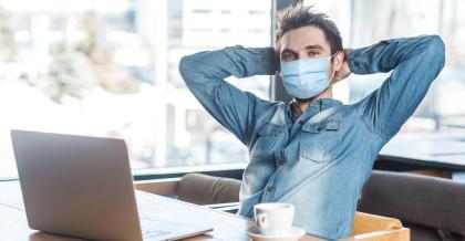 Ein Mitarbeiter einer Agentur sitzt mit Maske in einem Café an seinem Laptop.