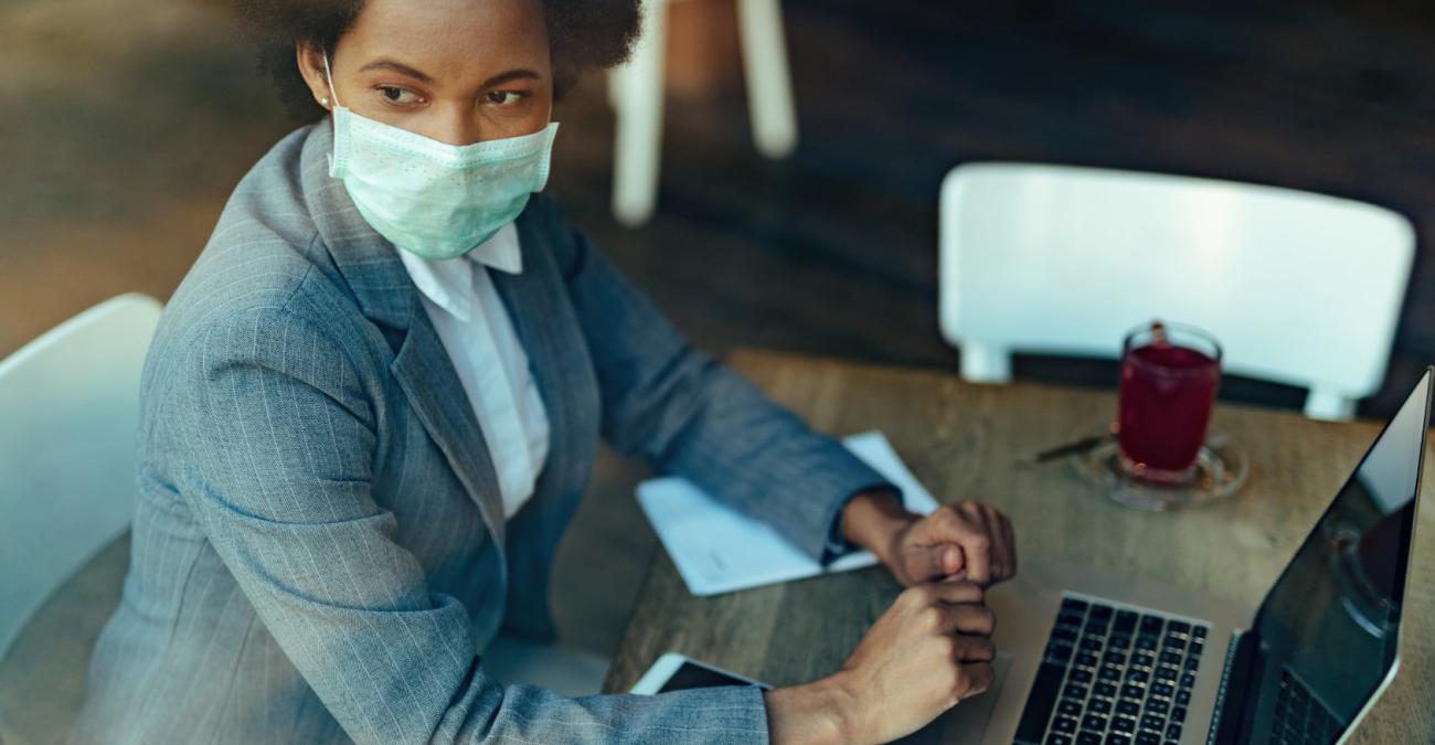 Digitale Transformation in Zeiten von Corona Eine Mitarbeiterin sitzt im Café und arbeitet mit Maske an ihrem Laptop.