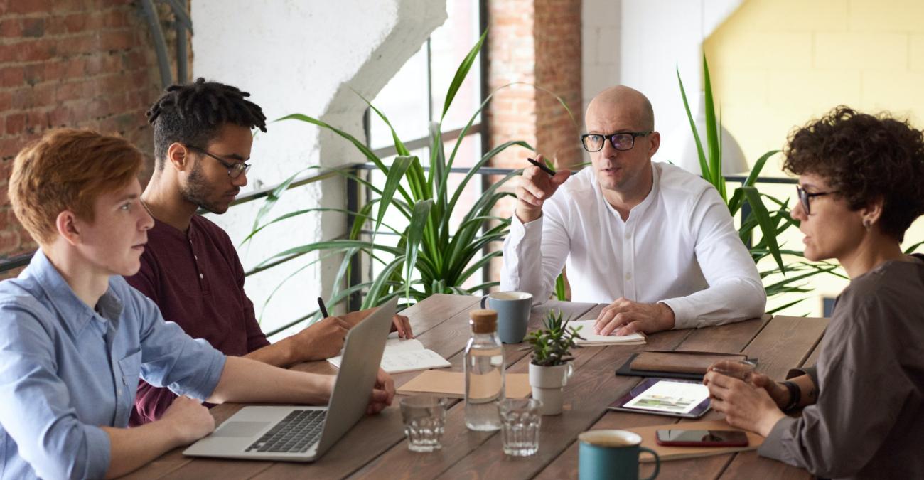 Menschen sitzen zusammen an einem Schreibtisch und haben Laptops und Tablets dabei und diskutieren