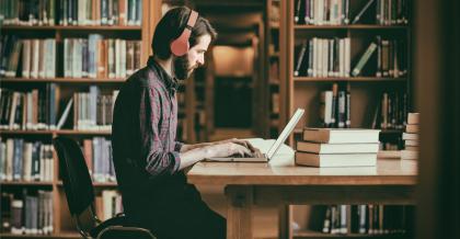 Junger Fachinformatiker sitzt in Bibliothek. Er arbeitet an seinem Notebook.