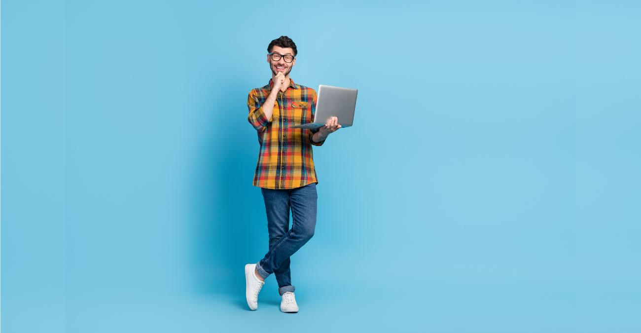 Junger Mann mit Notebook. Ein Anwendungsentwickler. Er steht dynamisch lächelnd vor blauer Wand.