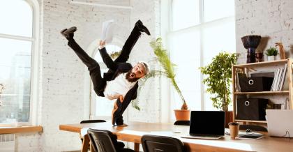 Junger Mann im Büro. Er macht lachend einen Handstand auf dem Schreibtisch. Er freut sich: Wir sind ISO 9001 zertifiziert.