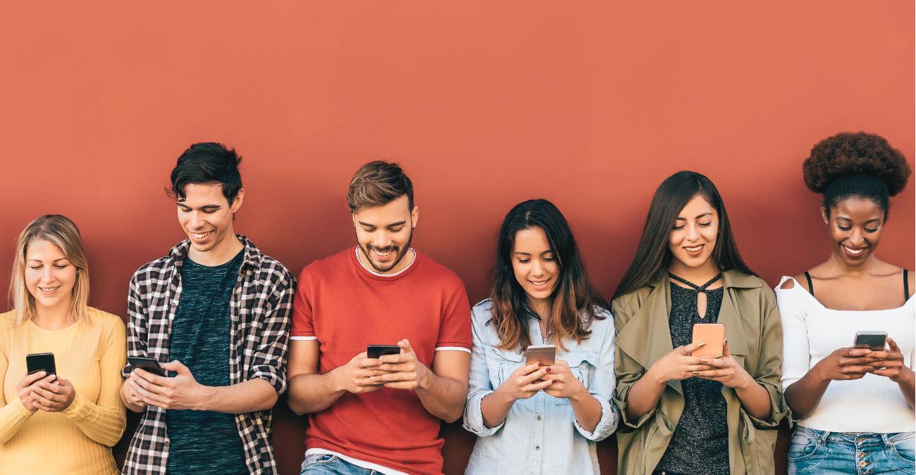 Social Tokens Sechs junge Menschen verschiedenen Geschlechts mit unterschiedlicher Hautfarbe stehen vor einer orangefarbenen Wand. Alle blicken auf ihr Smartphone und wollen Social Tokens kaufen.