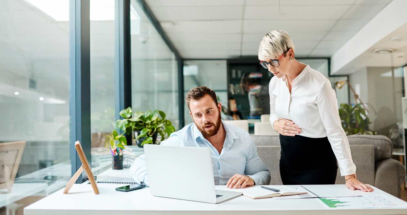 Ein Webentwickler sitzt im Büro an seinem Notebook. Seine Chefin steht neben ihm und berät ihn ber der Website-Performance-Optimierung.