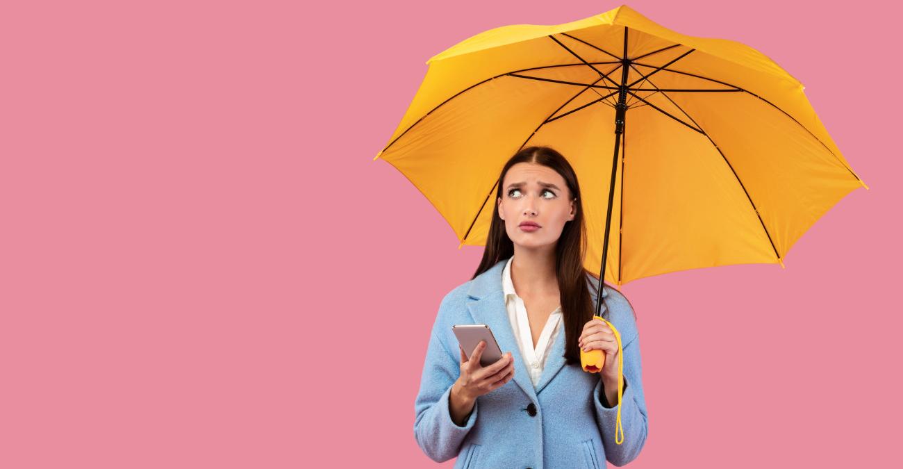 Eine junge Frau steht mit einem Regenschirm vor einer einfarbigen Wand. Sie hat ihr Handy in der Hand und blickt skeptisch zum Himmel. Ein Symbolbild für Preventive Maintenance.