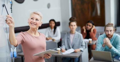 Eine fröhliche Lehrerin steht vor einer Klasse Erwachsener. Die erwachsenen Schüler gucken konzentriert. Sie machen eine Schulung: IT-Sicherheit.