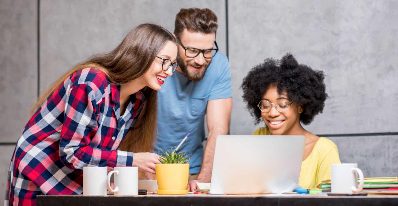 Ein junges dynamisches Team steht zusammen im Büro. Sie blicken auf den Bildschirm eines Notebooks und reden über CMS Hosting.