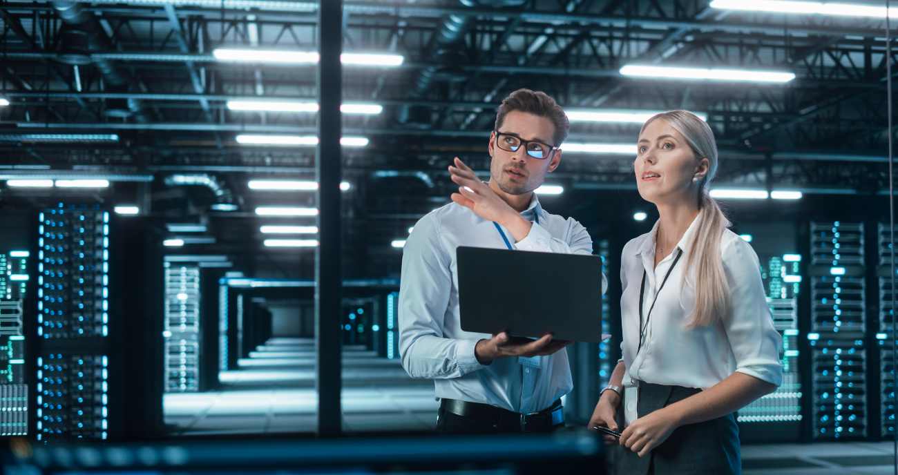 Ein dynamische Team aus Mann und Frau steht mit Notebook im Serverraum. Sie betreiben IT-Infrastruktur-Optimierung