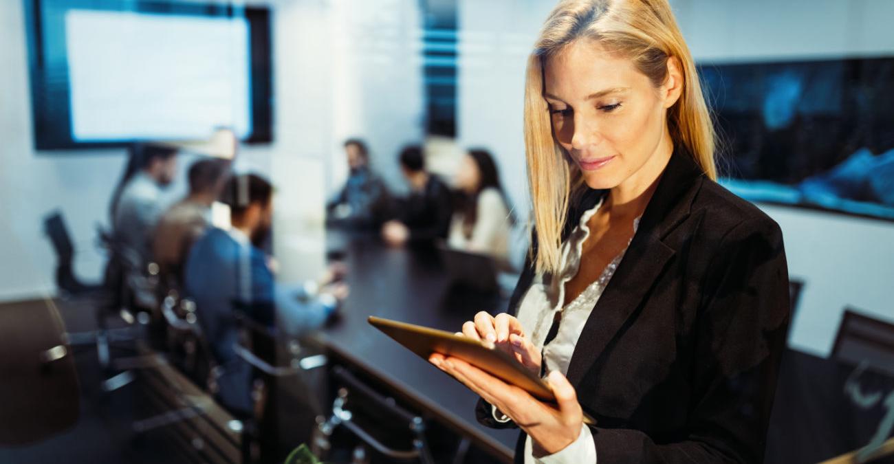 Eine junge Frau steht mit ihrem Tablet im Büro. Sie betreibt IT-Infrastruktur-Management. Im Hintergrund haben ihre Kollegen ein Meeting.