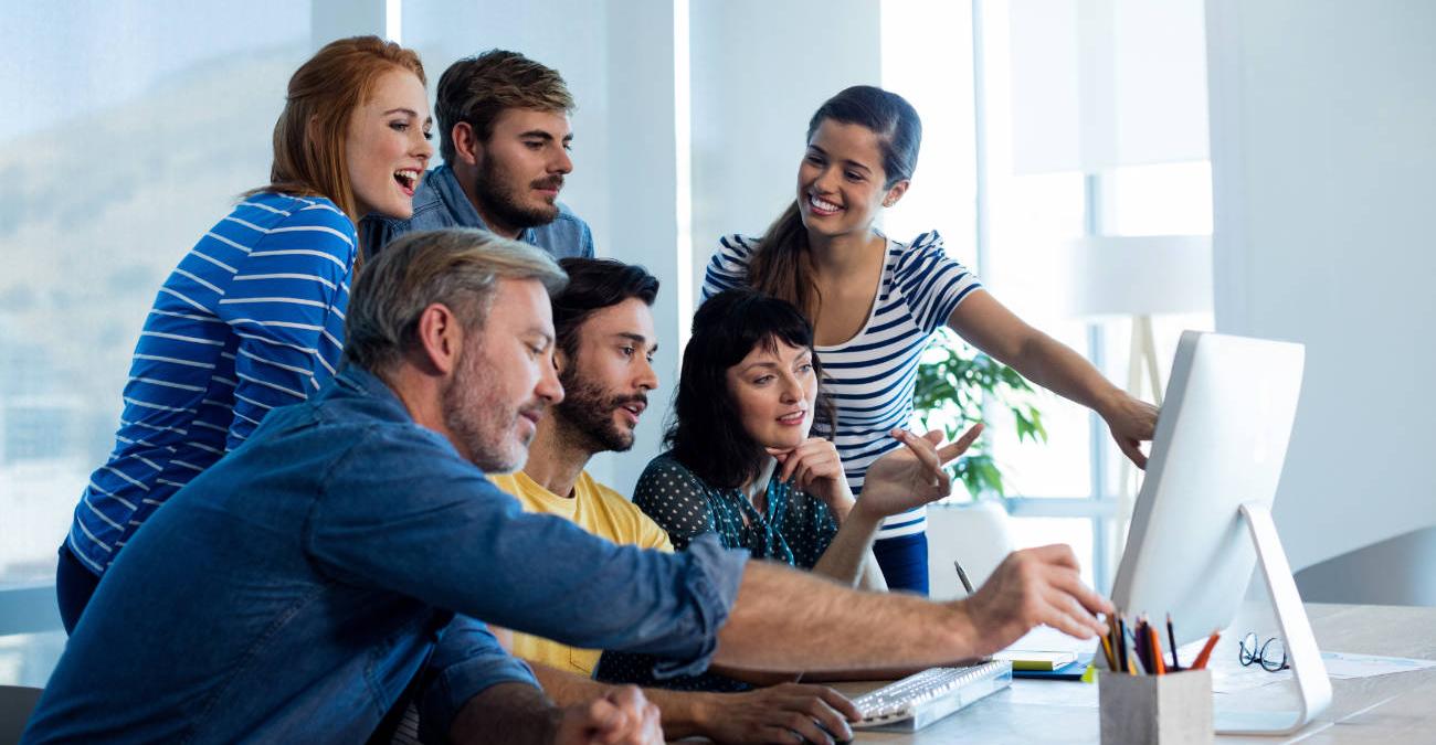 Ein helles und modernes Büro. Dynamische Mitarbeiter haben sich um einen PC versammelt und betreiben IT-Notfallmanagement.