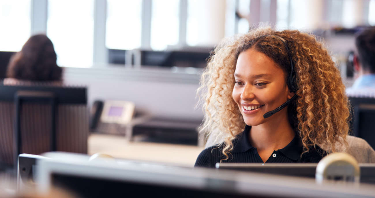 Eine junge Servicekraft sitzt am Rechner. Sie trägt ein Headset und lächelt freundlich. Sie berät einen Kunden zu den IT Support Levels.