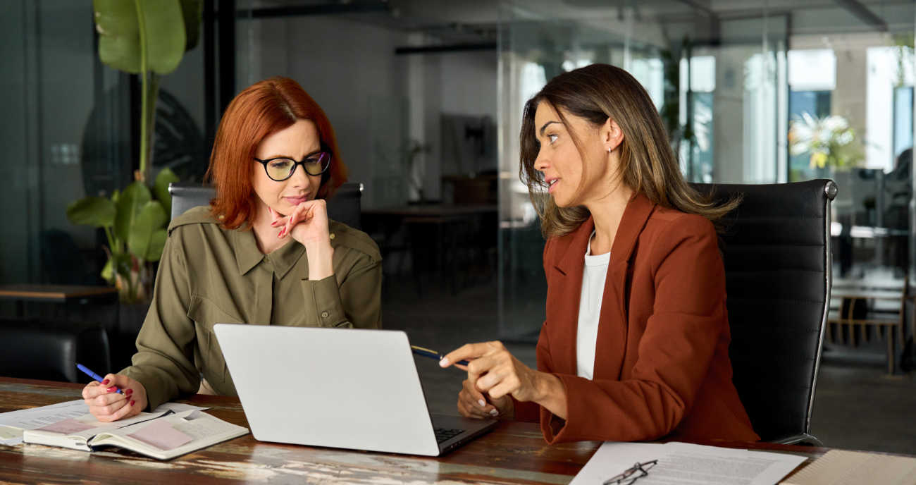 Zwei Frauen im Businessoutfit sitzen im Büro. Sie schauen auf ein Notebook und debattieren über die neue Software für Unternehmer.