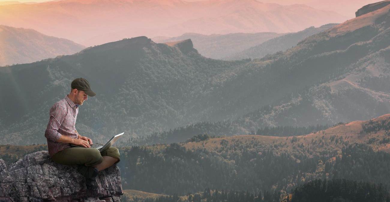 Barrierefreies Webdesign Ein Mann sitzt auf einem Felsen vor einer riesigen weiten Gebirgskette. Er hat ein Notebook auf dem Schoß. Die Weite der Landschaft ist ein Symbol für barrierefreies Webdesign.