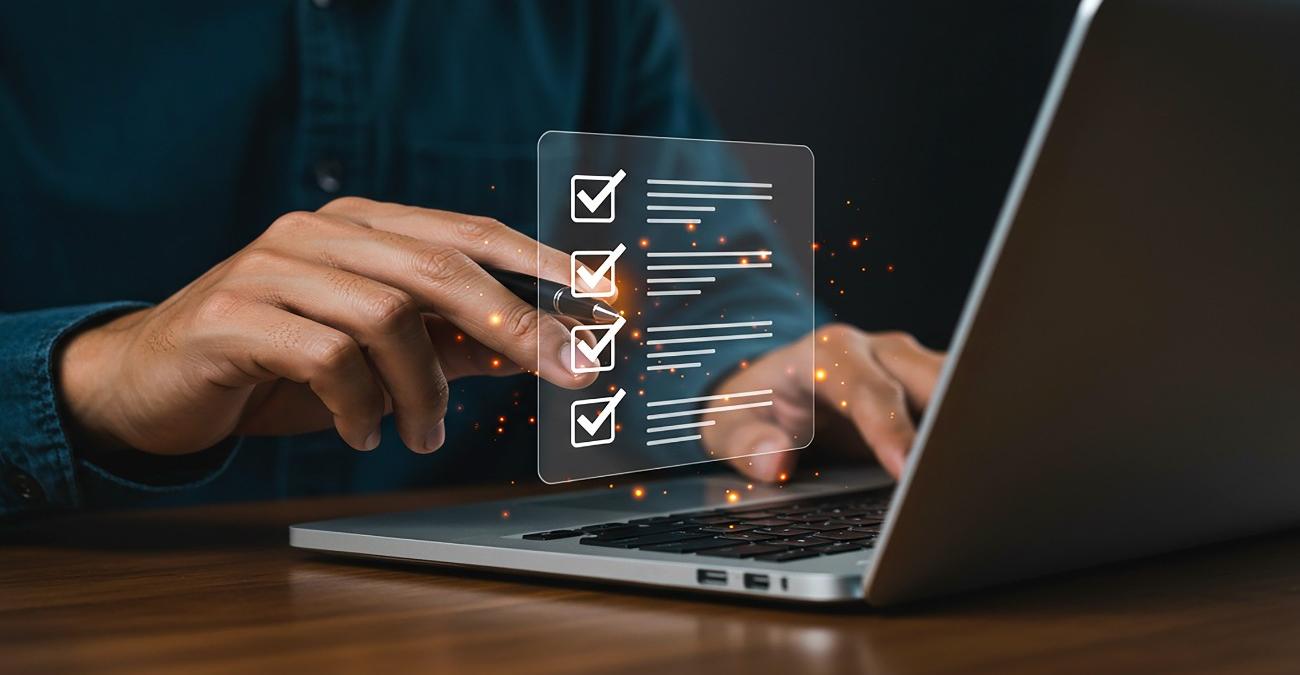 Detailaufnahme. Ein aufgeklappter Laptop. Daneben die Hand eines Mannes. Sie hält einen Stift. Er kreuzt damit Häkchen in Kästchen auf einer in die Luft projizierten Liste ab. Die Liste für die ISO 27001 Zertifizierung.
