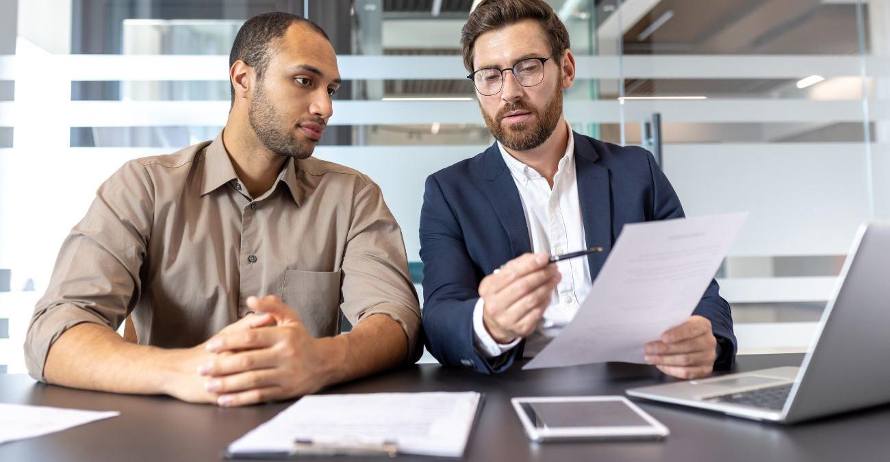 Zwei Männer in Business-Kleidung. Sie setzen mit einer Liste vor einem Notebook und beraten über eine ISO 27001 Zertifizierung.