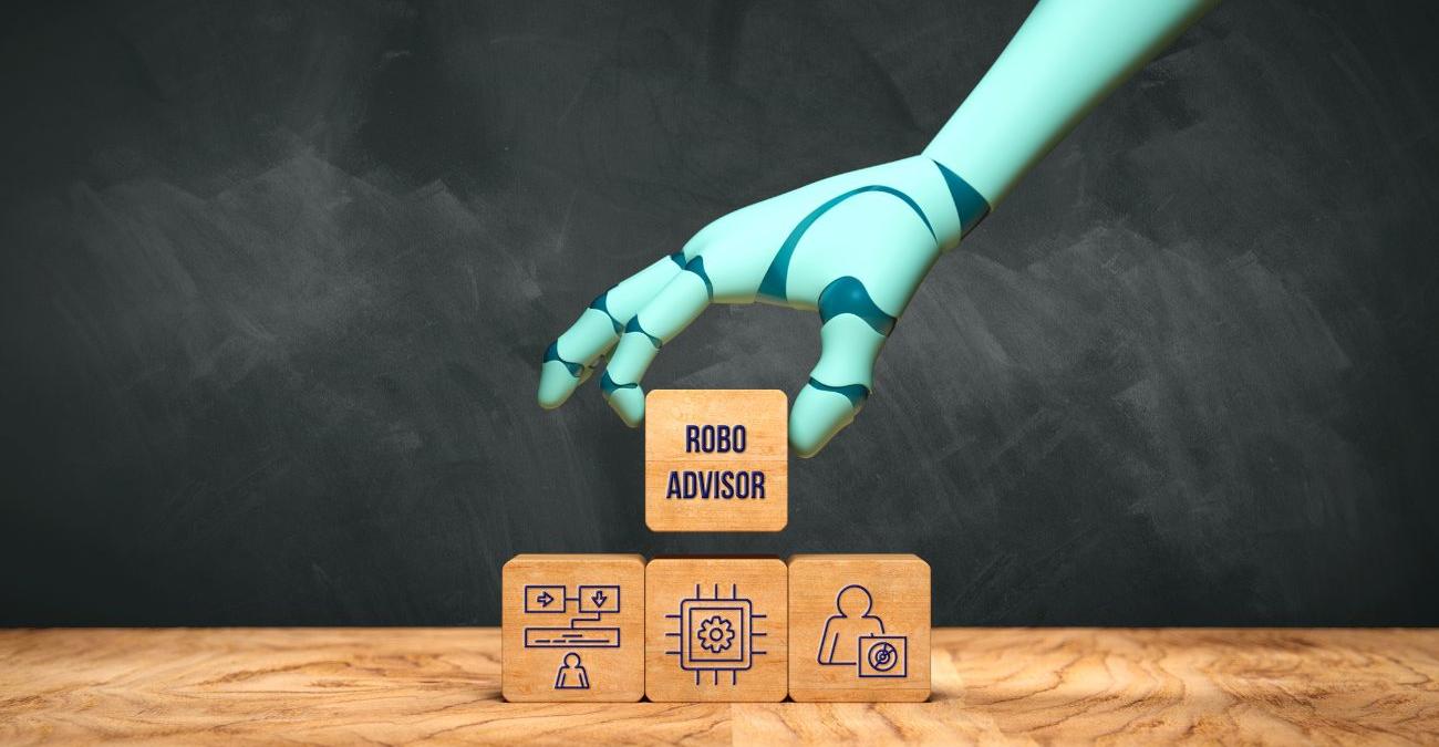 Mehrere Holzklötze liegen auf einem Tisch vor einer Schiefertafel. Eine Roboterhand hebt den oberen Klotz hoch. Die Aufschrift darauf lautet Robo Advisor.