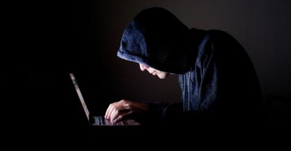 Halbdunkel. Ein Mann mit Kapuze sitzt über ein Notebook gebeugt. Er ist ein Hacker, der gerade einen NPM Supply-Chain-Angriff ausführt.