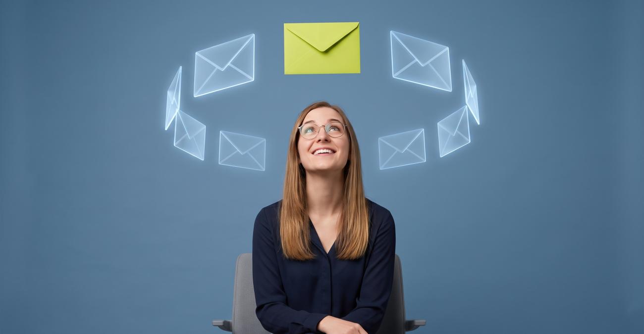 Eine junge Frau mit Brille sitzt auf einem Bürostuhl und blickt zu einem gelbgrünen Briefumschlag, der über ihr in einem Kreis aus durchscheinenden digitalen Umschlägen schwebt. Sinnbild für die interaktive E-Mail, in der die Handlung direkt in der Nachricht stattfindet.