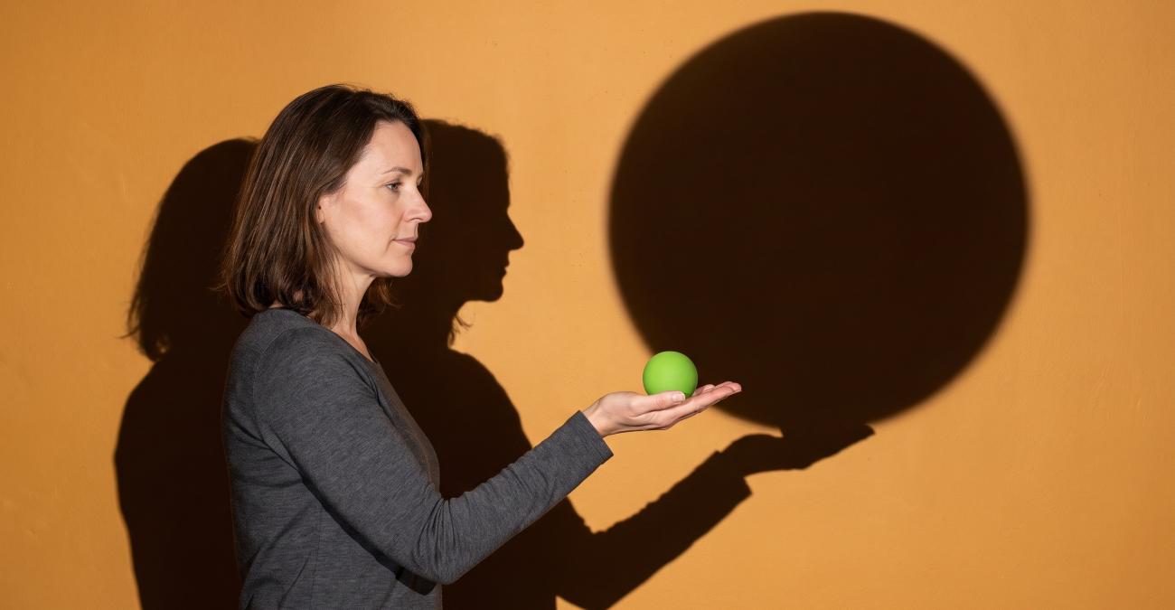 Eine Frau hält eine kleine gelbgrüne Kugel in der offenen Hand, deren Schatten die ganze Wand füllt. Sinnbild für die Kosten der Softwarewartung, die größer sind als das Sichtbare.