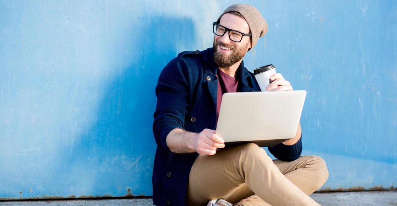 Sonnenschein. Draußen sitzt vor einer blauen Wand ein dynamischer Programmierer lässig auf dem Boden. Er trinkt Kaffee und arbeitzet an seinem Notebook mit dem Drupal CMS.