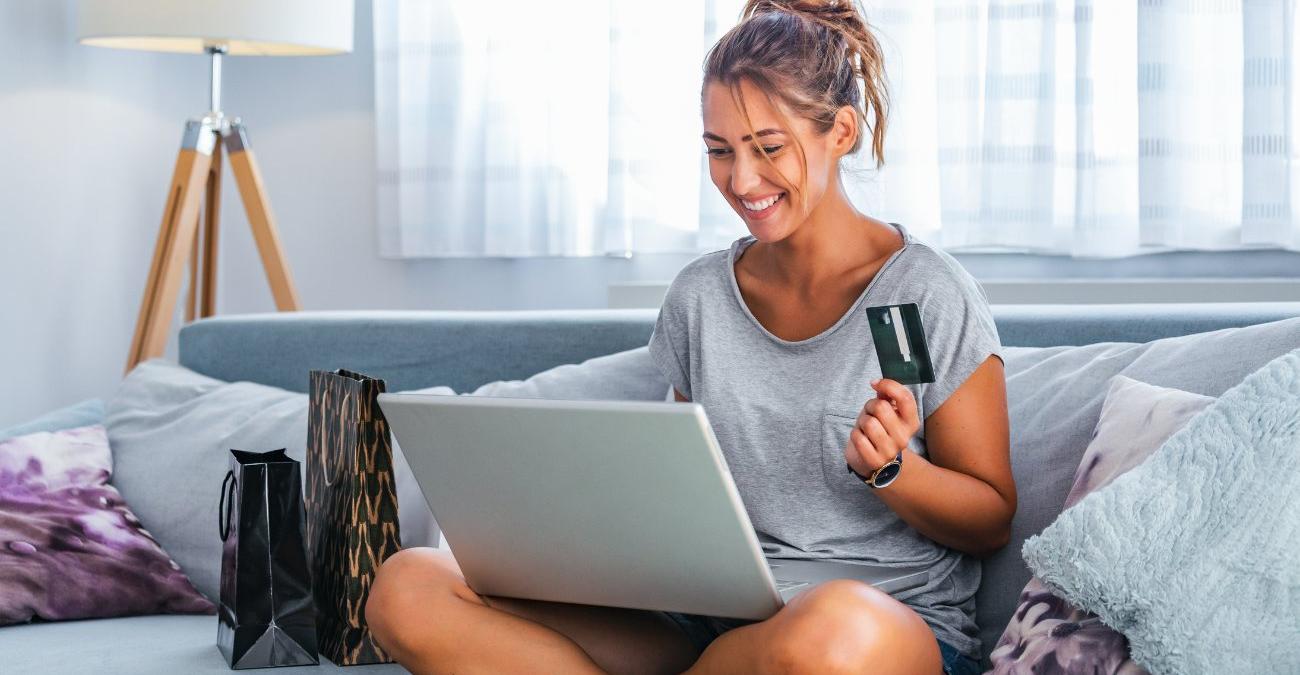 Ein helles Wohnzimmer. Eine junge Frau sitzt auf dem Sofa. Sie hat ihr Notebook auf dem Schoß und ihre Kreditkarte in der Hand. Gut gelaunt nutzt sie E-Commerce Software.