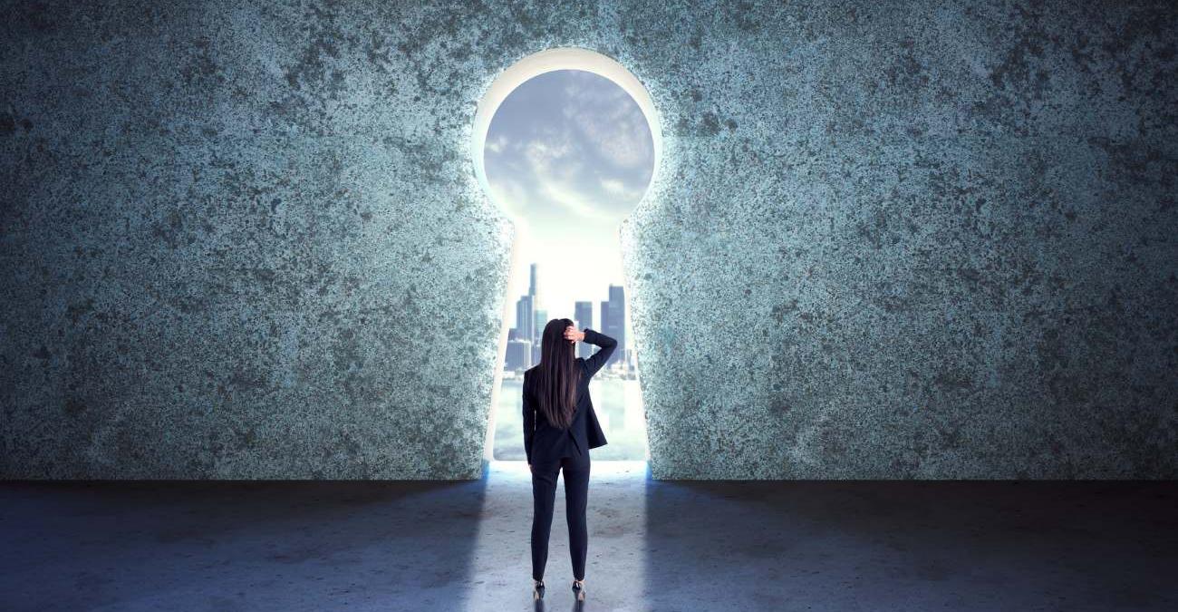 Eine Frau im Businessoutfit steht vor einer großen Felswand, in der ein Loch in Form eines riesigen Schlüssellochs klafft. Sie blickt durch das Schlüsselloch hinaus auf eine Großstadt. Ein Symbolbild für Vendor-lock-in.