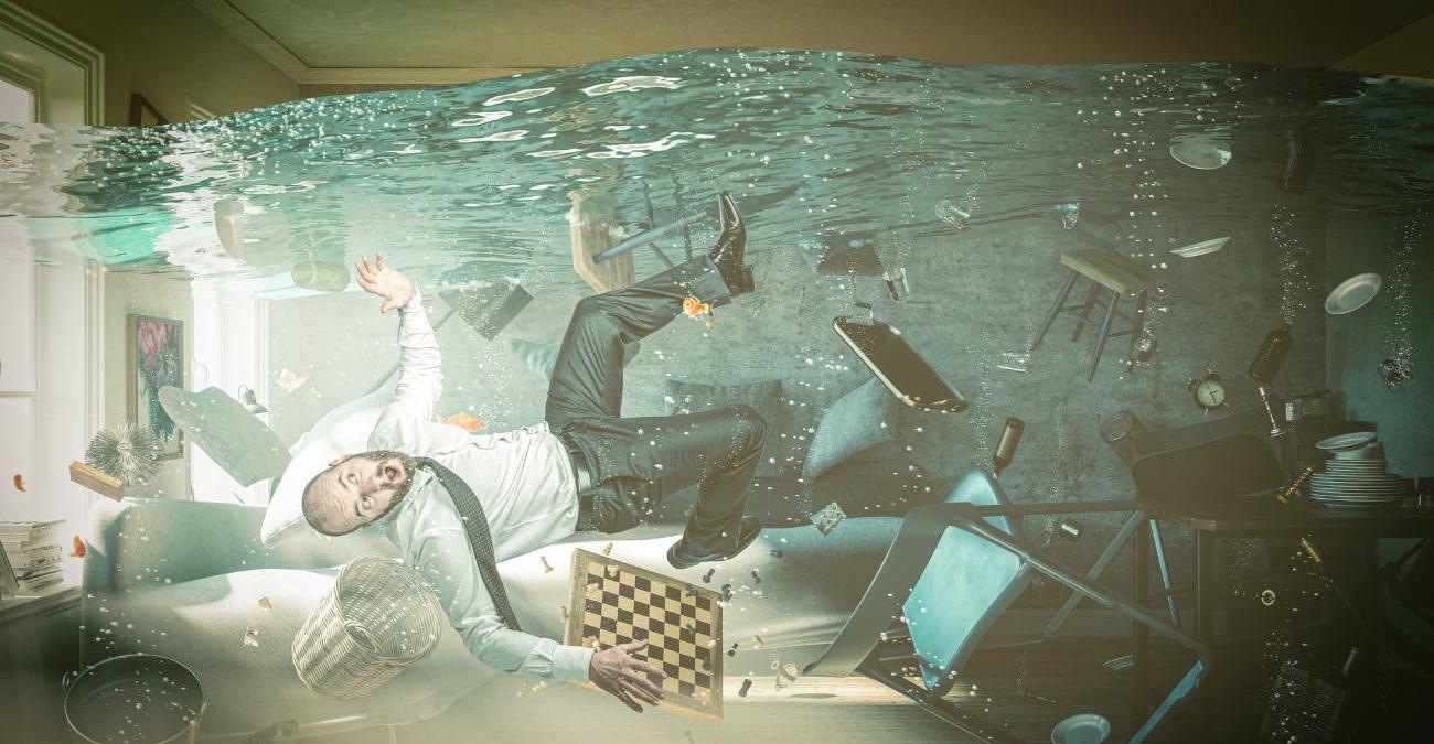 Ein Wohnzimmer steht völlig unter Wasser. Zwischen im Wasser schwebenden Möbeln treibt überrascht ein Mann. Ein eugenzwinkerndes Bild, das zeigt, dass krisenmanagement-software Hilfe für jederman sein kann.