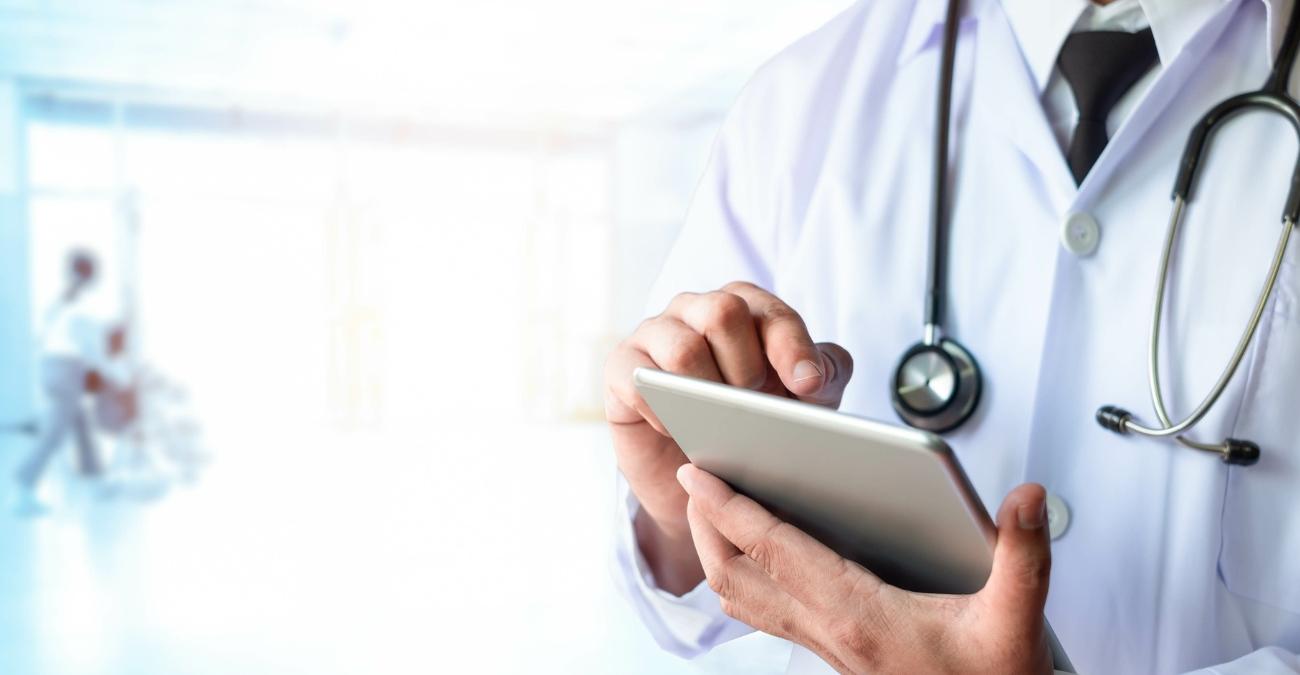 Arzt mit Stethoskop nutzt ein Tablet zur digitalen Diagnose und Informationsrecherche während einer Patientenuntersuchung, Klinikumgebung im Hintergrund – Sinnbild für  Health-IT im medizinischen Versorgungsalltag.