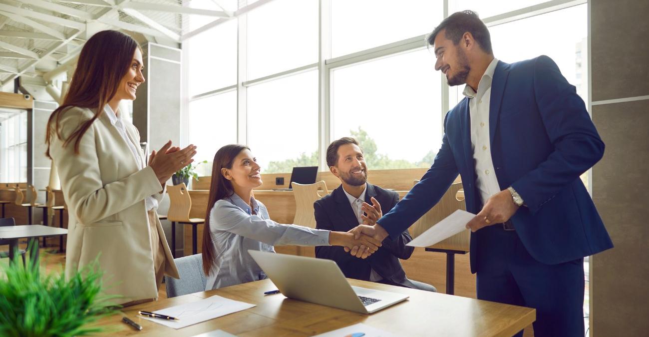 Geschäftspartner geben sich nach einem erfolgreichen Vertragsabschluss die Hand, während Kolleginnen und Kollegen im Büro applaudieren – Sinnbild für den gelungenen      EVB-Vertrag in der öffentlichen IT-Beschaffung.