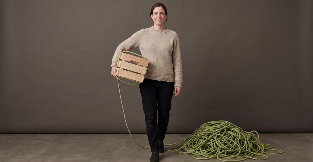 Eine Frau trägt eine kleine Holzkiste, aus der eine gelbgrüne Schnur zu einem riesigen Knäuel am Boden führt. Sinnbild für die Pflichten aus dem Cyber Resilience Act.