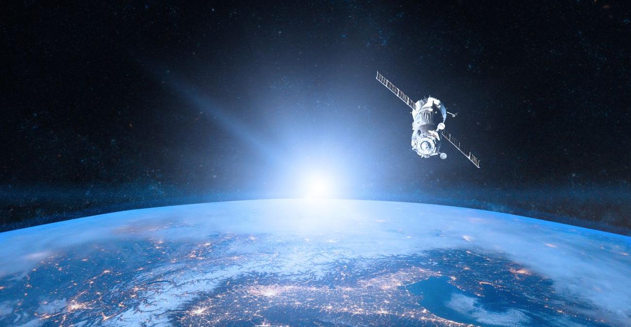 Die Erde vom Weltall betrachtet. Die Sonnenstrahlen bahnen sich ihren Weg daran vorbei. Ein Satellit für Erdbeobachtung schwebt darüber.