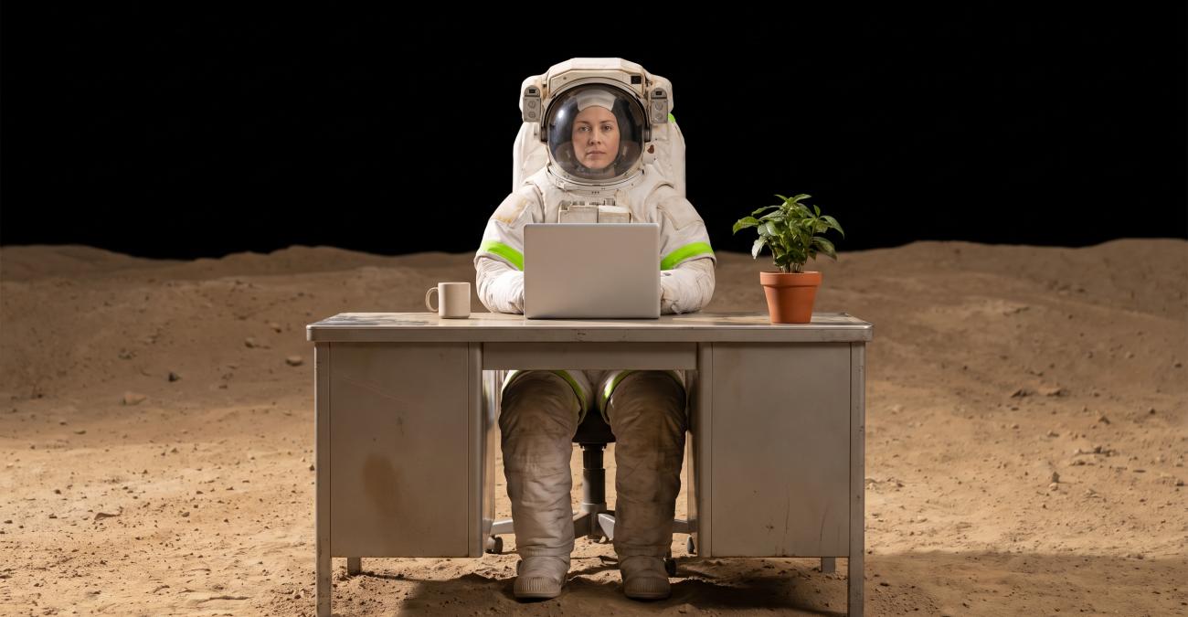 Eine Frau im Raumanzug sitzt an einem gewöhnlichen Büroschreibtisch auf einer staubigen Mondlandschaft unter schwarzem Himmel, auf dem Tisch Laptop, Tasse und Zimmerpflanze. Ein Sinnbild für eine Umgebung, die überallhin mitgenommen wird.