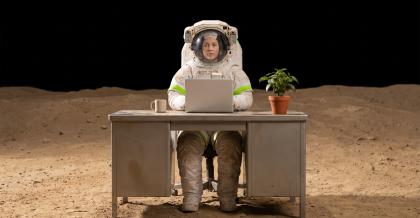 Eine Frau im Raumanzug sitzt an einem gewöhnlichen Büroschreibtisch auf einer staubigen Mondlandschaft unter schwarzem Himmel, auf dem Tisch Laptop, Tasse und Zimmerpflanze. Ein Sinnbild für eine Umgebung, die überallhin mitgenommen wird.