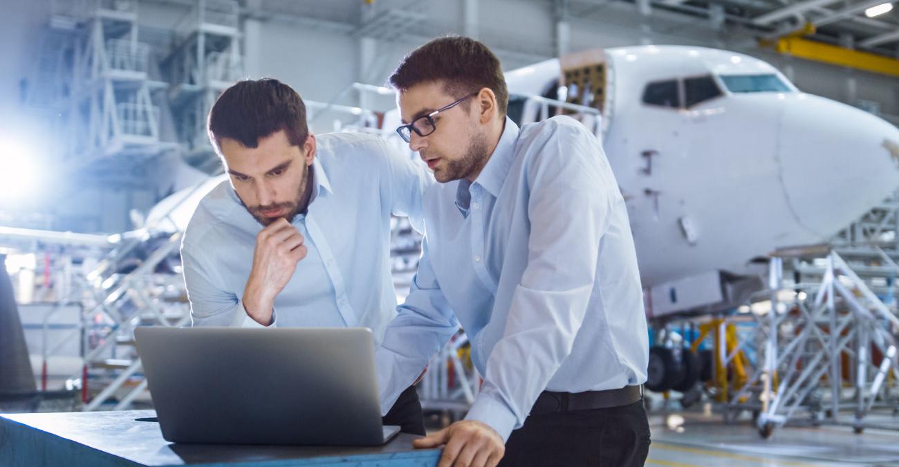 Zwei Männer stehen in einem kleinen Flugzeughanger. Sie sind über ein Notebook gebeugt und arbeiten mit Unternehmenssoftware.